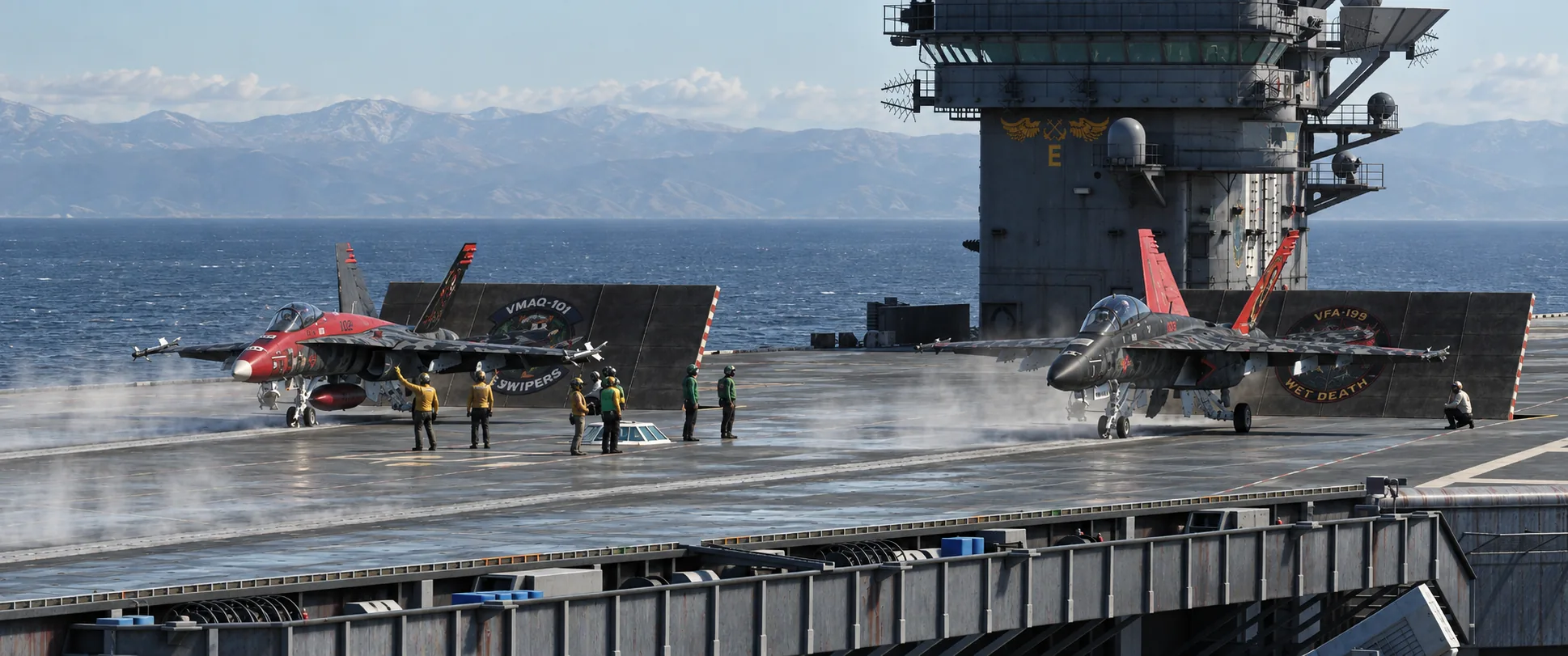 A VMAQ-101 Swipers Hornet and a VFA-199 Wet Death Super Hornet on the catapults with deck crew, the carrier island and mountains behind