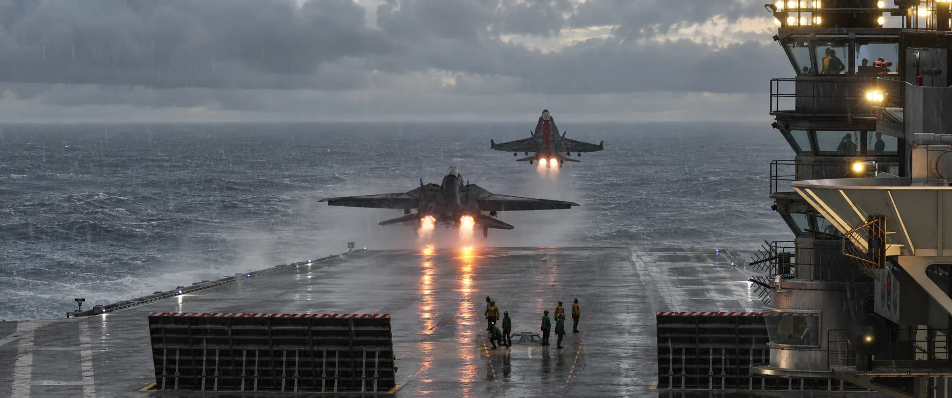 Two jets launching in afterburner off a rain-soaked carrier deck in a storm, deck crew and the island in the foreground